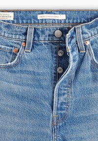 Vaqueros de mezclilla azul con un corte recto en los tobillos. Características incluyen remaches de cobre, un diseño clásico de cinco bolsillos y una textura desteñida.