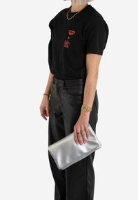Srebrna metaliczna kopertówka trzymana w ręku, noszona z czarnym t-shirtem i spodnie skórnymi o wysokim stanie, prezentująca minimalistyczny design.