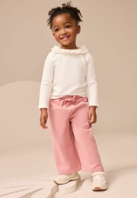 Una bambina sorridente che indossa una maglietta bianca con colletto arricciato, pantaloni rosa a gamba larga e sneaker bianche, sta in piedi su forme astratte color beige.