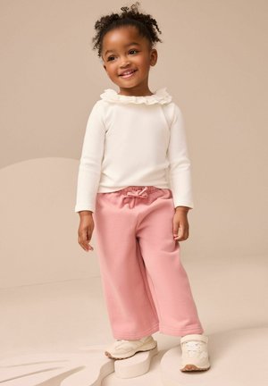 Lächelndes Kleinkind-Mädchen, das ein weißes Oberteil mit Rüsche, rosa weite Hosen und weiße Turnschuhe trägt und auf abstrakten beigen Formen steht.