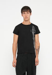 Czarny bawełniany t-shirt z krótkim rękawem, z białym abstrakcyjnym wzorem po lewej stronie. Model w ciemnych, kraciastych spodniach.