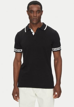 Uomo che indossa una polo nera a maniche corte con rifiniture bianche e testo sulle maniche, abbinata a pantaloni bianchi, in piedi davanti a uno sfondo semplice.