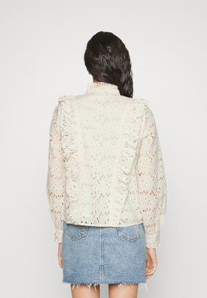 Woman with dark hair wearing a white lace blouse with ruffles and a light blue denim skirt, facing away from the camera.