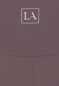 Paarse leggings met een subtiele geribbelde textuur, omrand met een wit rechthoekig logo met "LA," gepositioneerd bij de tailleband.