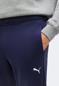 Personne portant un sweat gris et un pantalon de survêtement Puma bleu marine avec la main dans la poche, montrant le logo Puma sur le haut de la jambe.