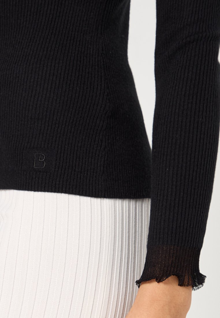 Czarny sweter prążkowany z długimi rękawami, z subtelną metką z logo. Rękawy mają przezroczysty, falbaniasty brzeg. Połączony z białymi prążkowanymi spodniami.