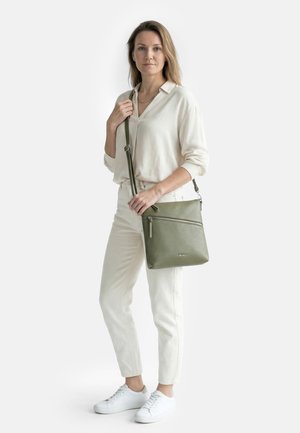 Mujer con conjunto blanco y zapatillas blancas, sosteniendo un bolso cruzado de cuero verde con detalle de cremallera.