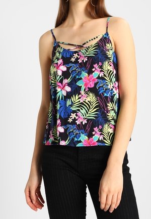 Mujer con un top negro sin mangas con un estampado floral tropical colorido y tirantes cruzados en el escote, combinado con pantalones negros.