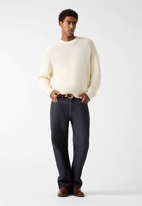 Pull en tricot crème, coupe ample, associé à un jean en denim bleu foncé. Ceinture en cuir marron avec boucle dorée, chaussures marron. Tenue simple et décontractée.