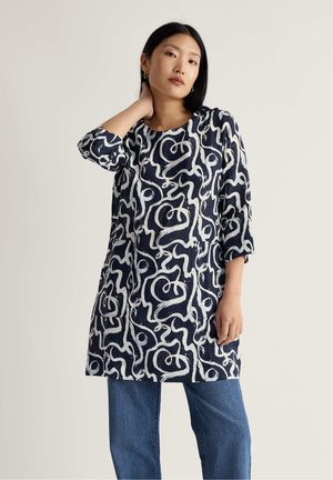 Moteris dėvi tamsiai mėlyną tuniką su balta abstrakčia vingiuota rašto ir trijų ketvirčių rankovėmis, derina su mėlynais džinsais, stovi priešais paprastą foną.