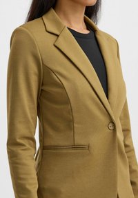 Olivově zelený šitý blazer s klopami, s jedním knoflíkem a detailními postranními kapsami. Nosený přes černý top z texturované tkaniny.