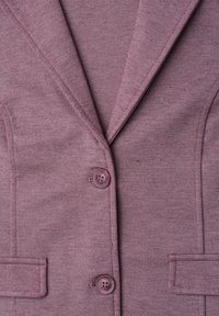 Gros plan d'un blazer mauve texturé avec deux boutons et deux poches à rabat à l'avant.