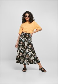 Gul oversized t-skjorte kombinert med et svart maxiskjørt med blomsterdesign og tropiske mønstre; sorte sandaler fullfører antrekket.