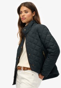 Veste matelassée bleu foncé avec un col montant, des accents marron, des accessoires dorés et une poche zippée, portée avec un pull crème et un jean.