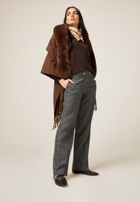 Brown poncho with faux fur trim, layered over a dark sweater and gray plaid wide-leg trousers. Black slip-on shoes complete the look.