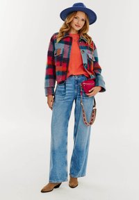 Farebná károvaná vlnená bunda s modrými, červenými a tealovými odtieňmi, spárovaná s oranžovým svetrom a svetlými riflami s vysokým pásom. Hnedé lodičky.