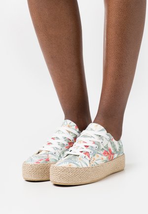 Pies con zapatillas blancas de plataforma con suelas de cuerda tejida y patrones florales coloridos sobre un fondo blanco liso.