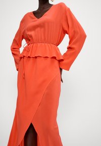 Langes orangenes Kleid aus leichtem Stoff mit V-Ausschnitt, gerafftem Taillenbereich, langen Ärmeln und seitlichem Schlitz.