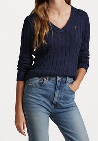 Mörkblå kabelstickad tröja med V-ringning, ribbade muddar och en liten logotyp på bröstet, tillsammans med ljusblå slitna jeans.