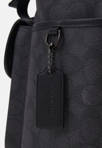 Zwarte leren tas met textuur, voorzien van een metalen ring en een label dat met een klein kettingtje is bevestigd, met reliëf merknaam op het label.