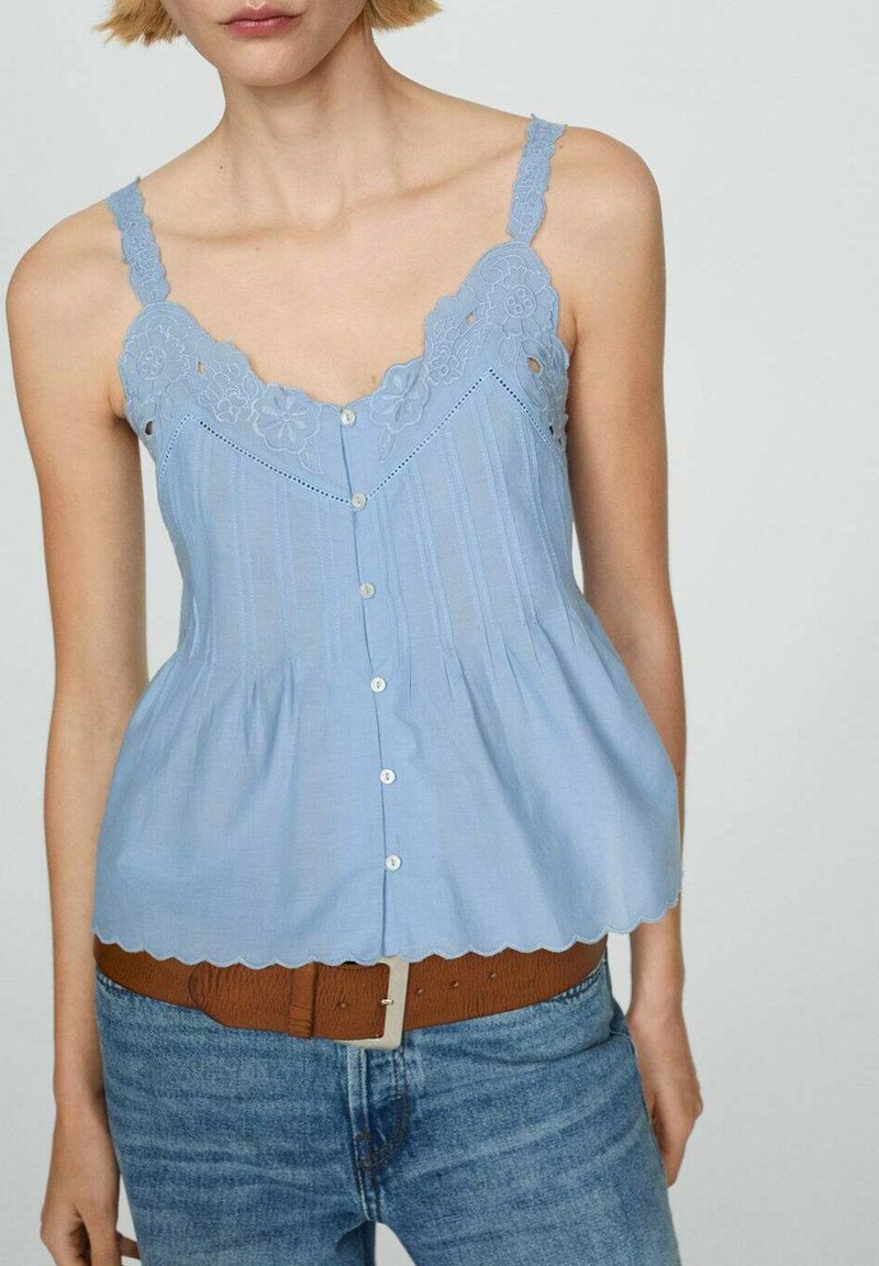 Jasnoniebieska bezrękawnik z koronkowymi detalami, pionowymi paskami, zapięciem na guziki i falbanastym brzegiem; zestawiona z brązowym paskiem i dżinsami.