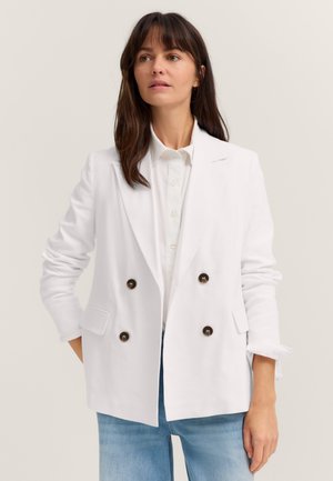 Frau mit braunen Haaren, die einen weißen, zweireihigen Blazer, ein weißes Hemd mit Knöpfen und hellblaue Jeans vor einem einfarbigen Hintergrund trägt.