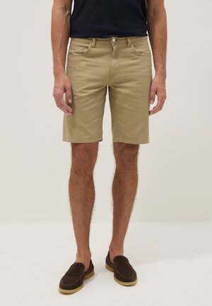 Mann trägt beige kniehohe Shorts, dunkle Loafers und ein schwarzes Hemd, steht vor einem schlichten hellen Hintergrund.