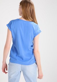 Top azul de manga corta en tejido ligero, de corte holgado, con escote redondo y puños enrollados, combinado con jeans azul claros.