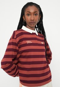 Burgundska in rjavo prugasta polo majica z dolgimi rokavi in belim ovratnikom, ki vsebuje vezena besedila "Atletska oblačila" in logotip na prsih.