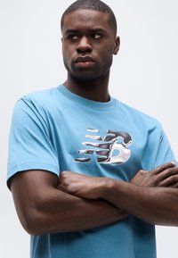 Νέος άνδρας με σταυρωμένα χέρια φοράει κοντομάνικη μπλούζα σε ανοιχτό γαλάζιο χρώμα με μεταλλικό ασημί λογότυπο "NB" σε απλό λευκό φόντο.