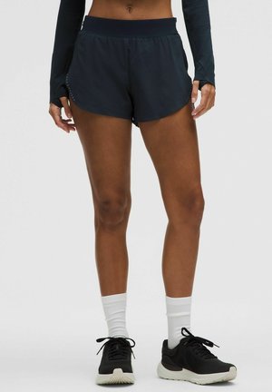 Frau trägt schwarze Sportshorts, langärmeliges Oberteil, weiße Socken und schwarze Laufschuhe und steht vor einem schlichten Hintergrund.