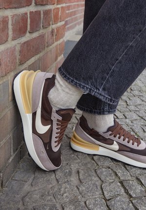 Osoba nosząca brązowe i beżowe buty Nike, beżowe skarpetki oraz ciemne dżinsy, stojąca na kamiennej kostce brukowej przy ścianie z cegły.