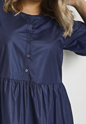 Vrouw die een marineblauwe jurk met knopen en een geplooide taille draagt, getoond van nek tot halverwege de dij, terwijl ze met één hand haar haar opstopt.