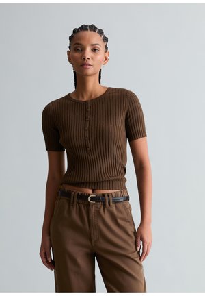 Młoda kobieta z warkoczami, ubrana w brązową, prążkowaną, zapinaną na guziki bluzkę z krótkim rękawem oraz brązowe spodnie z wysokim stanem i czarnym paskiem.