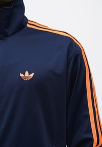 Μπουφάν σε ναυτικό μπλε με πορτοκαλί λεπτομέρειες τριών ρίγες στα μανίκια και πορτοκαλί λογότυπο Adidas trefoil στο στήθος.