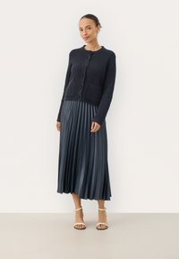 Cardigan en tricot bleu marine avec fermeture à boutons et deux poches, associé à une jupe midi plissée bleu marine. Le modèle porte des sandales à brides beiges.