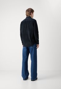 Camicia di velluto a coste nera con colletto a punta, polsini con bottone e vestibilità dritta, abbinata a pantaloni in denim blu larghi e scarpe nere.