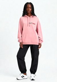 Felpa rosa con tasca frontale, con grafica 'SWEET heart'. Abbinata a pantaloni cargo neri e sneakers bianche.