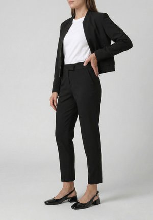 Kobieta ubrana w czarne, dopasowane spodnie, czarną krótką kurtkę na białej koszulce i czarne niskie szpilki typu slingback, stojąca na szarym tle.