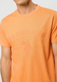 Trumpas rankovės oranžinis medvilninis marškinėlis su blukusiu polo žaidėjo grafiku ir tekstu "U.S. POLO ASSN." bei apvaliu kaklo dizainu.