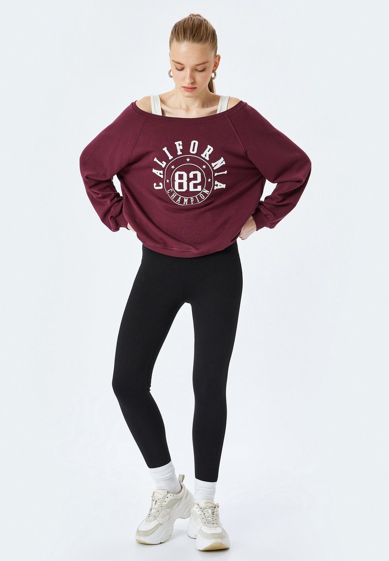 Bordeaux oversized sweatshirt met witte "California 82" grafiek, gecombineerd met zwarte leggings en beige sneakers. Eenvoudig ontwerp met geribbelde manchetten.