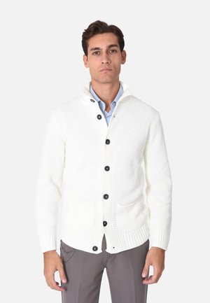 Cardigan bianco lavorato a maglia realizzato in tessuto testurizzato, con cappuccio, due tasche frontali e bottoni neri. Abbinato a una camicia azzurra chiara.