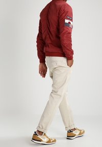 Czerwona bomberka z marszczonym tyłem i naszywką z amerykańską flagą na rękawie, zestawiona z kremowymi spodniami i brązowymi sneakersami Nike.