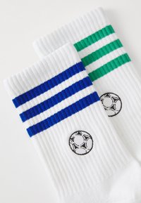 Baltas zeķes ar zilu un zaļu svītru rakstu un melnu izšūtu futbola bumbu logotipu. Ir ribota tekstūra un kontrastējoši krāsas akcenti.