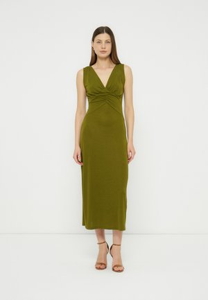 Kvinde iført ærmeløs olivengrøn midi-kjole med rynket V-udskæring og brune ankelremssko, stående mod en ensfarvet hvid baggrund.