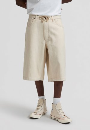 Mand iført beige, teksturerede knælange shorts med reb-bælte, hvide sokker og beige høje sneakers foran en ensfarvet baggrund.