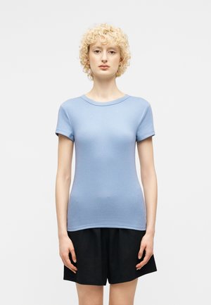 Frau mit kurzen lockigen blonden Haaren, die ein hellblaues, kurzärmeliges, eng anliegendes Hemd und schwarze Shorts trägt, steht vor einem schlichten Hintergrund.