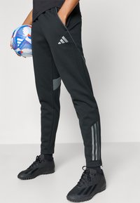 Pantaloni atletici negri cu accente gri, croială strâmtă, cu trei dungi verticale pe lateral și buzunare. Este ținută o minge de fotbal cu model.
