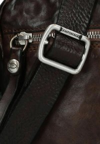 Sezione di borsa in pelle marrone scuro con una fibbia in metallo argentato, una tracolla con texture a contrasto e una cerniera in metallo con un dettagli embossato del logo.