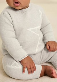 Väike laps kannab heledat halli kudumit ja pükse, istub pehmel kreemikollasel pinnal, käed ja paljad jalad on nähtavad.